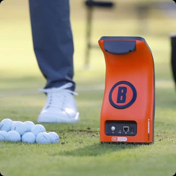 Bushnell Launch Pro im orangen Gehäuse auf dem Rasen der Driving Range, daneben ein Spieler und ein Haufen Übungsbälle