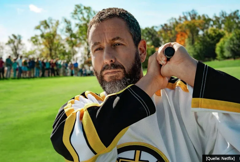 Adam Sandler als Happy Gilmore in einem Boston-Bruins-Eishockey-Trikot, den Golfschläger wie einen Hockey-Stick haltend auf dem Fairway, mit Zuschauerkulisse im Hintergrund (Quelle: Netflix)