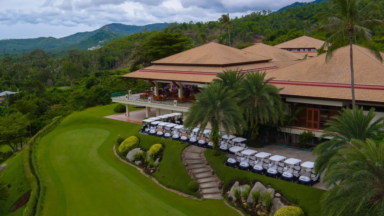 Luftaufnahme des Clubhauses des Santiburi Samui Country Club mit pavillonartigen Dächern, aufgereihten Golfcarts und Blick über die bewaldeten Berge Koh Samuis