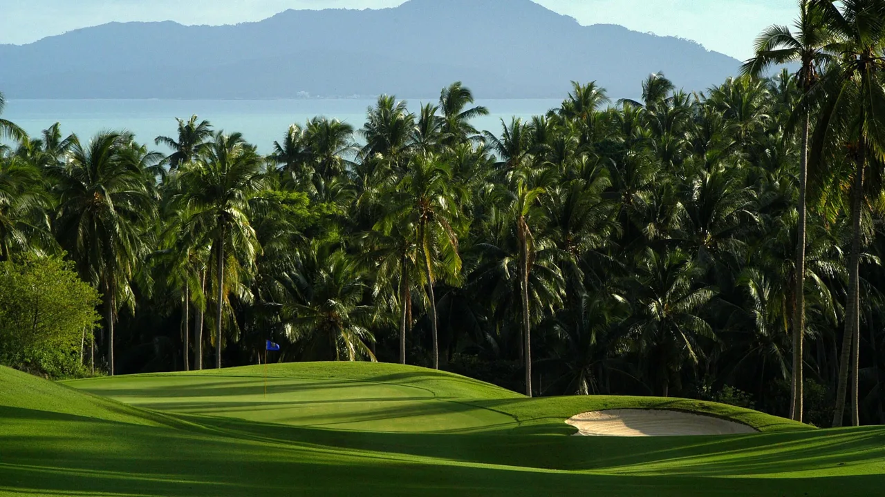 Ein Grün des Santiburi Samui Country Club mit Bunker, gesäumt von Kokospalmen, dahinter der Golf von Thailand und die Berge der Nachbarinsel